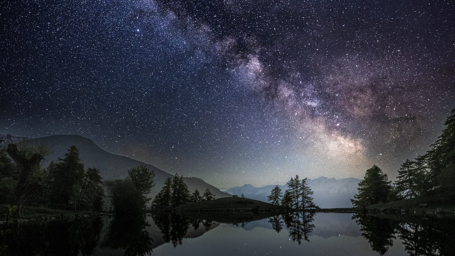 Die Milchstraße spiegelt sich in einem stillen Bergsee mit Baumsilhouetten